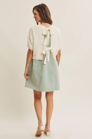 A woman shown from the back wearing a short cream knit cardigan with a large bow detail at the back and a light sage green skirt, paired with beige wedge sandals, against a plain cream studio background.
