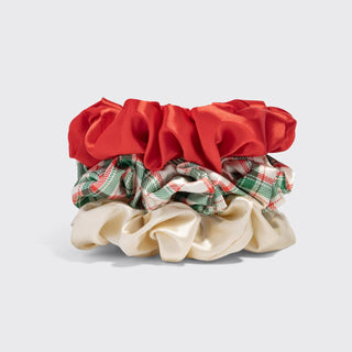 Three satin hair scrunchies stacked together on a light gray background: a bright red scrunchie on top, a green and red plaid scrunchie in the middle, and a cream-colored scrunchie on the bottom.