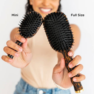 Person holding a mini and full-size hairbrush with text labels on a plain background