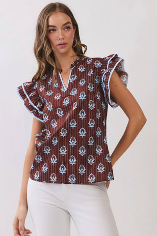 A woman modeling a short-sleeve blouse with flutter sleeves, a split V-neckline, and an allover white floral block print on a dark reddish-brown base, paired with white pants against a light gray studio background.