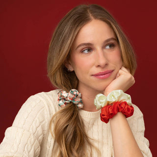 A woman with long light brown hair wearing three satin hair scrunchies in red, cream, and a floral pattern, posed against a solid dark red background.