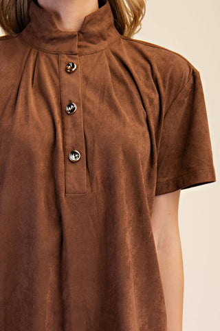 A close-up of a person wearing a brown short-sleeve mock-neck top with a buttoned placket and textured fabric, against a plain light beige background.