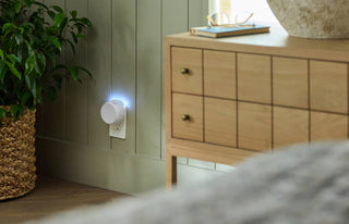 A white cylindrical smart plug or night light glowing with a soft blue light, plugged into a wall in a styled bedroom or living space next to a wooden cabinet and potted plant.