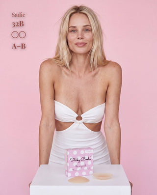 A blonde model wearing a white cutout bandeau dress stands against a pink background, with a pink box of Tidy Buns silicone pasties and two beige silicone pasties displayed on a white table in the foreground. Text on the top left reads 'Sadie 32B OO A-B'.