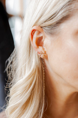 A close-up of a woman's ear wearing a delicate gold ear cuff with a long dangling chain earring, set against her blonde hair.