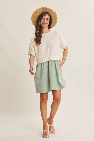 A woman modeling a cream short-sleeve knit top and a sage green skirt, styled with a straw hat and white strappy sandals against a light beige studio background.