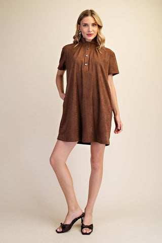 A woman modeling a brown short-sleeve mini dress with a high gathered neckline and decorative buttons, paired with dark brown heeled sandals, against a solid beige studio background.