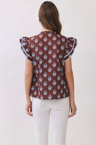 A woman viewed from the back wearing a brown ruffled blouse with a white floral block print and white pants, set against a plain off-white background.