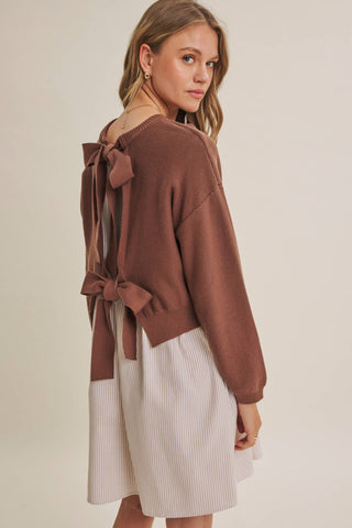 A woman shown from the back wearing a brown knit sweater with a large bow detail on the back and a light cream pleated skirt, set against a plain beige studio background.
