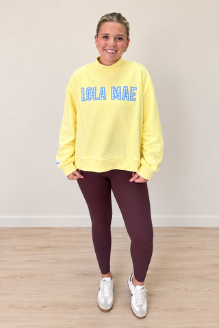 A woman modeling a pale yellow sweatshirt with 'LOLA MAE' printed in blue block letters across the chest, paired with dark burgundy leggings and white sneakers, standing against a neutral beige wall and light wood floor.