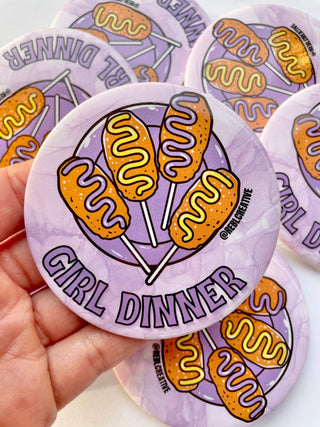 Sticker with corn dogs and 'Girl Dinner' text on a purple background, held by a hand.