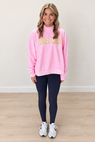 A woman modeling a pink sweatshirt with 'LOLA MIAE' printed in yellow and white across the chest, paired with dark leggings and white sneakers, standing against a neutral beige wall and light wood floor.