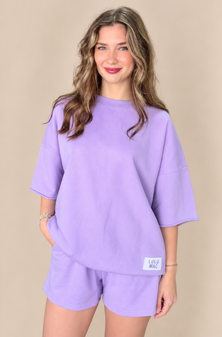 A woman modeling a matching lavender lounge set consisting of an oversized short-sleeve top and shorts, featuring a small white brand label on the bottom hem, set against a solid beige background.