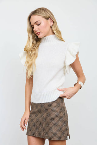 A blonde woman wearing a white sleeveless knit sweater with ruffled shoulders and a brown plaid mini skirt, posing against a light gray studio background.