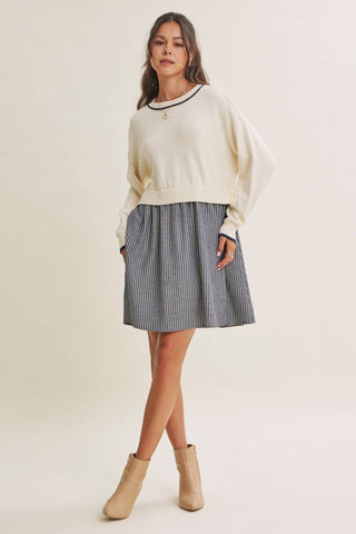 A woman modeling a cream knit cropped sweater with black trim, a blue and white vertically striped pleated mini skirt, and beige ankle boots, set against a plain cream studio background.