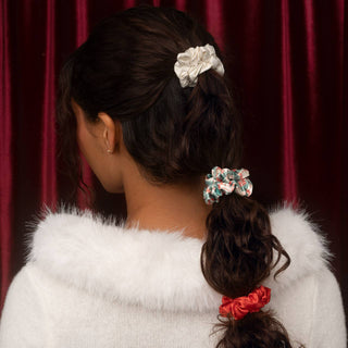 A woman shown from the back wearing three fabric hair scrunchies in white, multicolor floral, and red, styled in her dark brown hair against a dark red curtain background.
