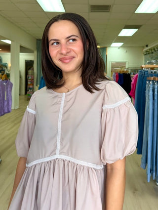 A woman with dark shoulder-length hair wearing a light pink puff-sleeve dress with white trim, standing inside a clothing boutique with racks of garments in the background.