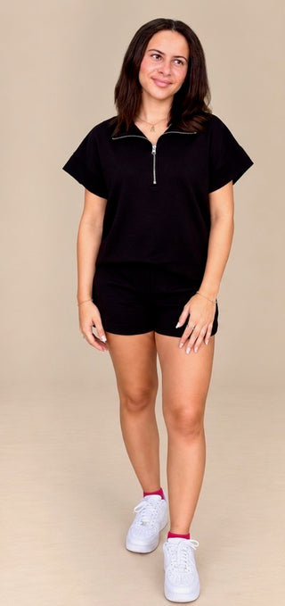 A woman modeling a black short-sleeve half-zip pullover top with matching black shorts, styled with white sneakers, against a solid beige studio background.