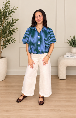 A woman modeling a blue and black vertically striped short-sleeve button-up shirt with white wide-leg pants and black lace-up flats, standing in a bright room with a light wood floor, white wall paneling, and potted plants.