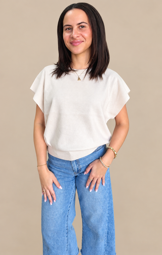 A woman modeling a cream-colored short-sleeve knit top with ruffled edges, paired with blue jeans, against a solid beige background.