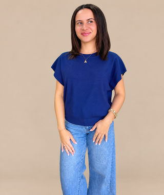 A woman modeling a royal blue short-sleeve top with wide, draped sleeves and light-wash blue jeans, standing against a solid beige studio background.