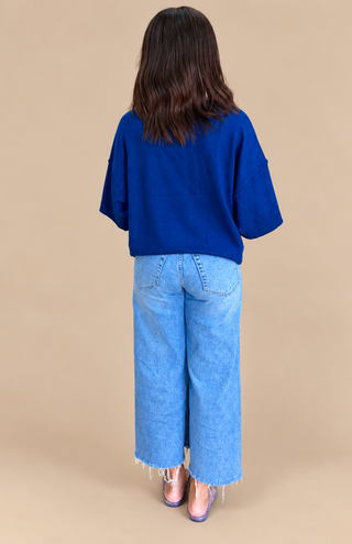 A woman shown from the back wearing a vibrant royal blue short-sleeve top and light wash wide-leg denim jeans with frayed hems, standing against a solid beige studio background.