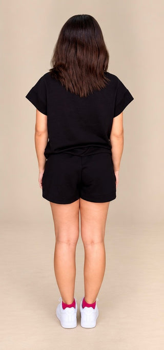 A child model shown from the back wearing a black short-sleeve t-shirt and matching black shorts, paired with white sneakers and pink socks, against a solid beige studio background.