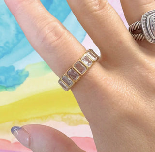 Gold ring with clear stones on a hand against a colorful watercolor background