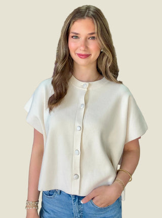 A woman wearing a cream-colored short-sleeve button-up top and blue jeans, standing inside a clothing boutique with shelves and garments visible in the background.