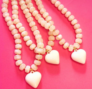 Three beaded necklaces with pale pink round beads and white heart-shaped pendants, arranged on a bright pink background.