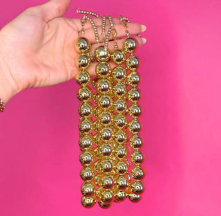 A hand holding five strands of shiny gold ball-bead fringe against a bright pink background.