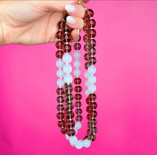 A hand with white manicured nails holds up a multi-strand beaded necklace featuring dark red, translucent brown, and opaque white beads separated by small gold spacer rings, set against a solid hot pink background.