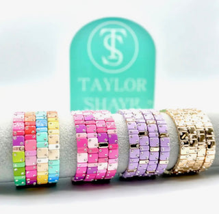 Colorful beaded bracelets on a white surface with a 'Taylor Shimmer' branded stand.