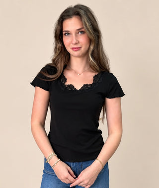 A woman modeling a black short-sleeve top with a black lace V-neck trim, paired with blue jeans, against a solid beige background.