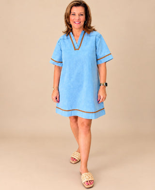 A woman modeling a light blue short-sleeve tunic-style dress with brown trim details, standing against a solid beige studio background.