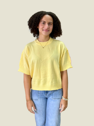 A woman with curly dark hair wearing a pale yellow short-sleeve knit sweater and light blue jeans, standing against a solid light beige background.
