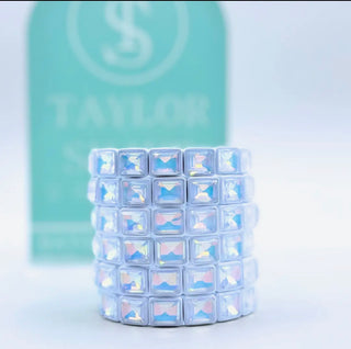 Stack of crystal-studded rings with a blurred 'Taylor' brand logo in the background.