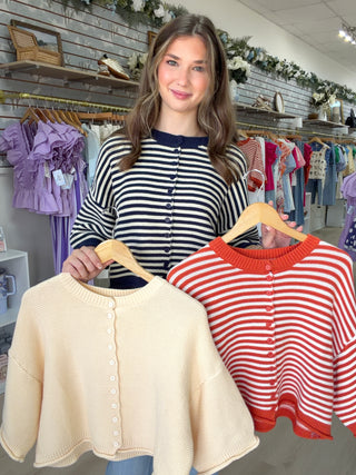 A woman in a clothing boutique holds up two striped cardigans on wooden hangers. The cardigan on the left is cream with a navy button placket, and the cardigan on the right has red and white horizontal stripes with a red collar and button placket.