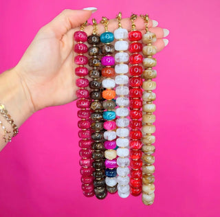 A hand holds six strands of beaded keychains against a bright pink background. The keychains feature round, polished beads in various colors including pink, brown, blue, white, orange, red, and beige, with gold-toned metal keychain hardware.