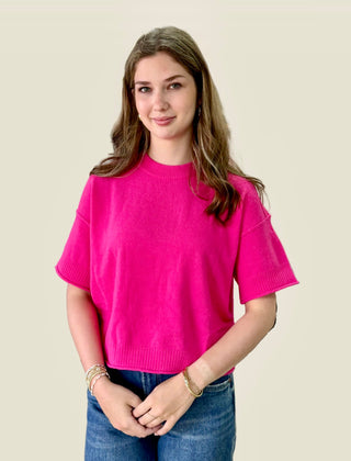 A young woman with long light brown hair wearing a bright pink short-sleeve knit sweater and blue jeans, standing against a solid light beige background.