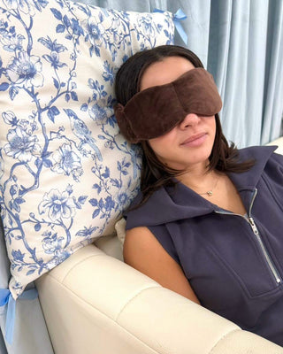 A woman reclining on a cream-colored sofa, wearing a brown plush eye mask and a dark purple sleeveless zip-up top, with a blue and white floral pillow behind her head.