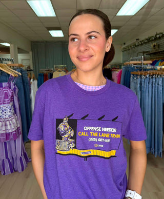 A woman wearing a purple graphic t-shirt with a cartoon train and the text 'OFFENSE NEEDED! CALL THE LANE TRAIN (225) GET-OUT' in a clothing store.