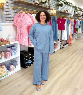 A woman stands in a boutique wearing a matching light blue and white striped lounge set, with clothing racks and shelves visible in the background.