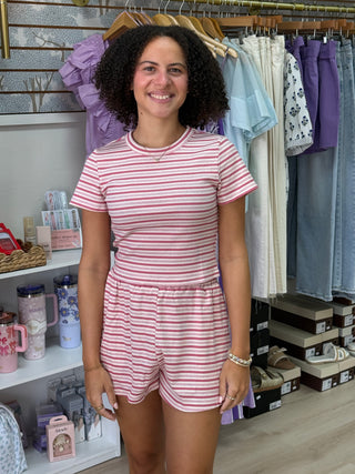 A smiling woman with curly dark hair stands in a boutique wearing a pink and white horizontally striped short-sleeve romper. Clothing racks and shelves are visible in the background.