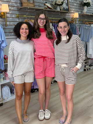 Three women standing together in a clothing boutique, wearing matching striped lounge sets. The set on the left is gray and white, the set in the center is pink and white, and the set on the right is brown and white.