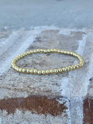 A thin beaded bracelet made of small metallic gold beads, resting on a textured gray stone surface.