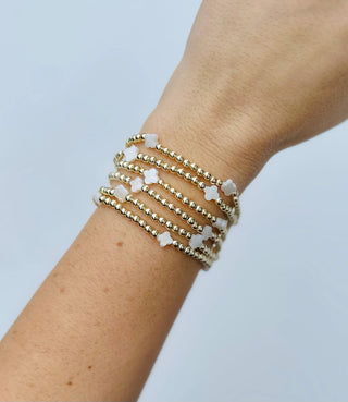 A close-up of a person's wrist wearing a stack of five delicate beaded bracelets. The bracelets feature small gold-toned beads and white flower-shaped charms, set against a clean, light blue background.