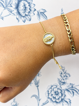 A close-up of a person's wrist wearing two gold bracelets: a delicate chain bracelet with a round white inlay and gold bird motif, and a thicker gold curb chain, against a white background with blue floral print.