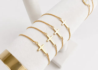 Four gold chain bracelets with small cross pendants displayed on a white cylindrical jewelry stand.