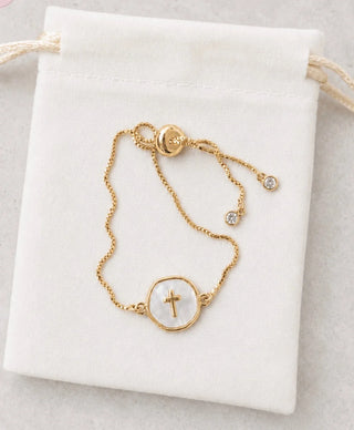 A delicate gold chain bracelet with a circular white shell pendant featuring a small gold cross, displayed on a white drawstring pouch.